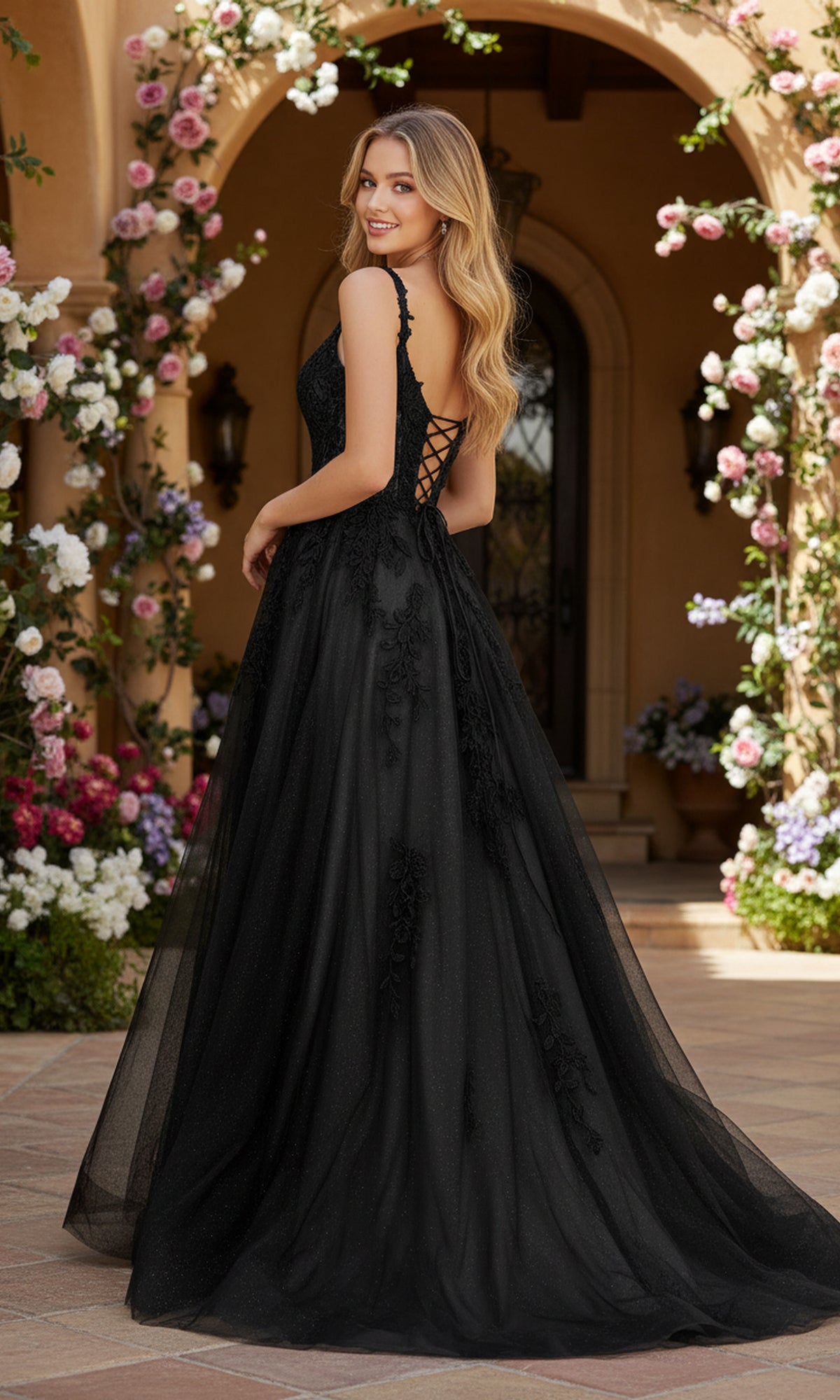 A woman smiles over her shoulder outdoors in front of a floral arch, wearing the PromGirl Exclusive 250261—a long black lace applique prom dress with a full skirt and floral detailing.